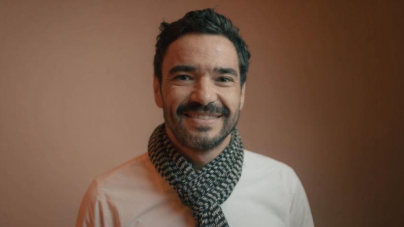 Homem de cabelos curtos e escuros sorri para a câmera, vestindo camiseta branca e cachecol preto com padrão branco, com fundo marrom uniforme.