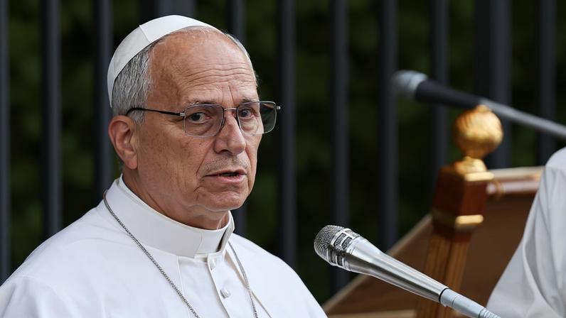 Homem vestido com vestes brancas e solidéu branco fala em microfone diante de púlpito de madeira, com grades e vegetação ao fundo.