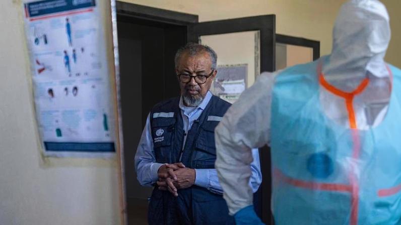 Homem de óculos e colete azul com logotipo da OMS observa ambiente interno de hospital. Ao fundo, outro homem e pessoa com equipamento de proteção individual azul e branco aparecem parcialmente.