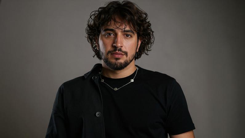 Homem jovem de cabelos cacheados e barba curta veste camiseta preta e jaqueta preta aberta, posando contra fundo branco neutro.