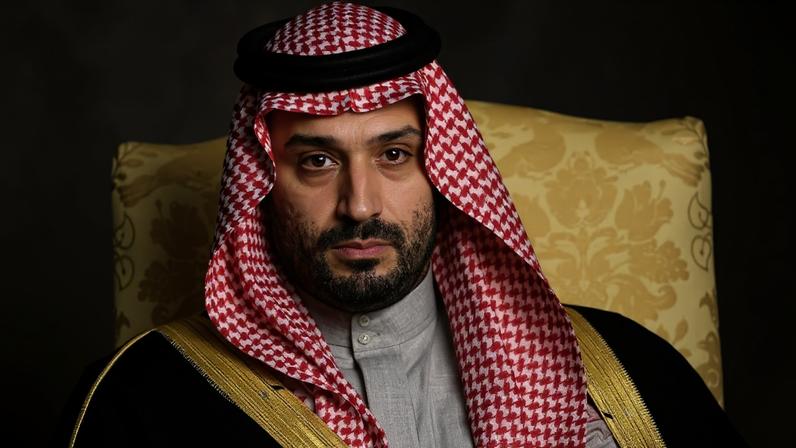 Homem árabe com barba curta veste traje tradicional com keffiyeh vermelho e branco preso por agal preto, e bisht preto com detalhes dourados, sentado em poltrona amarela com padrão floral.