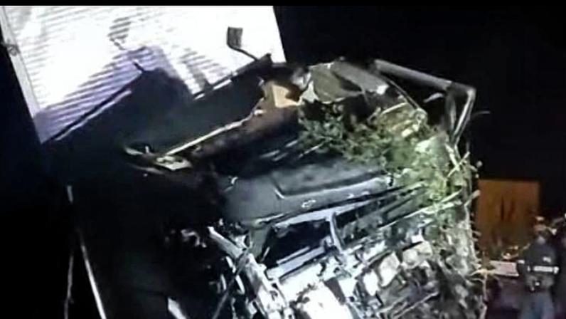 Caminhão tombado de lado à noite com a cabine amassada e parcialmente destruída. Vegetação está presa na estrutura do veículo. Iluminação artificial destaca os danos enquanto equipes de emergência estão próximas.
