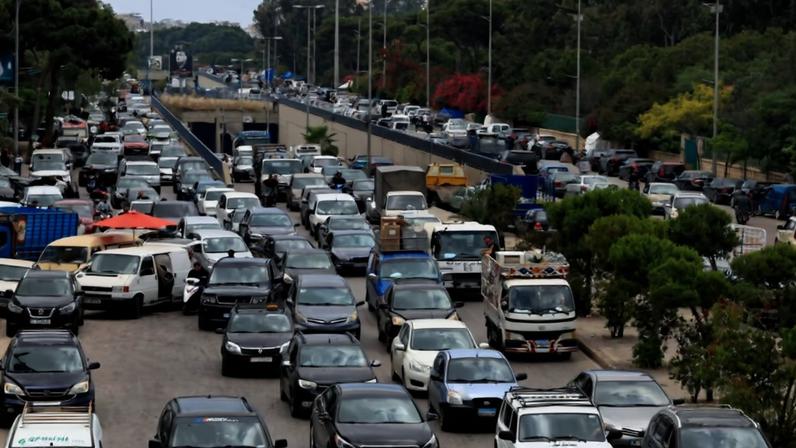 Congestionamento pesado em via urbana com várias faixas ocupadas por carros, caminhões e vans. Vegetação e árvores ao lado da pista, com viaduto ao fundo e céu nublado.