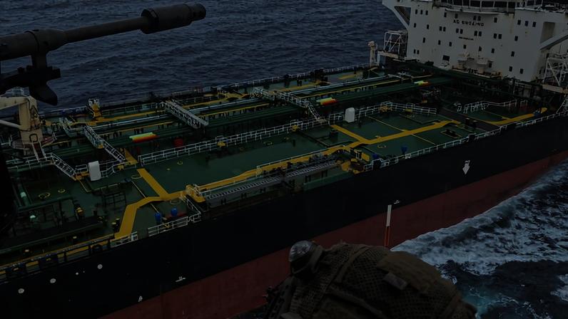 Militar com capacete e equipamento tático observa navio petroleiro no mar a partir de helicóptero. Navio tem casco preto e vermelho, com deck verde e tubulações amarelas.