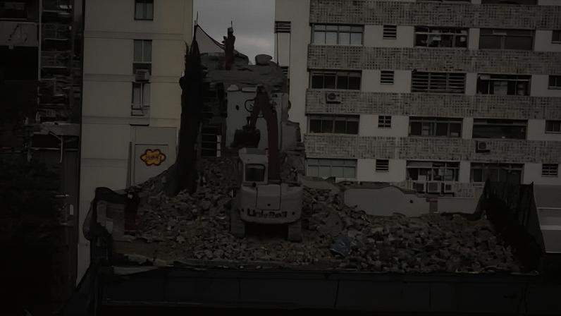 Máquina escavadeira realiza demolição no topo de um prédio cercado por outros edifícios residenciais altos. Entulho cobre o terraço enquanto a escavadeira opera.