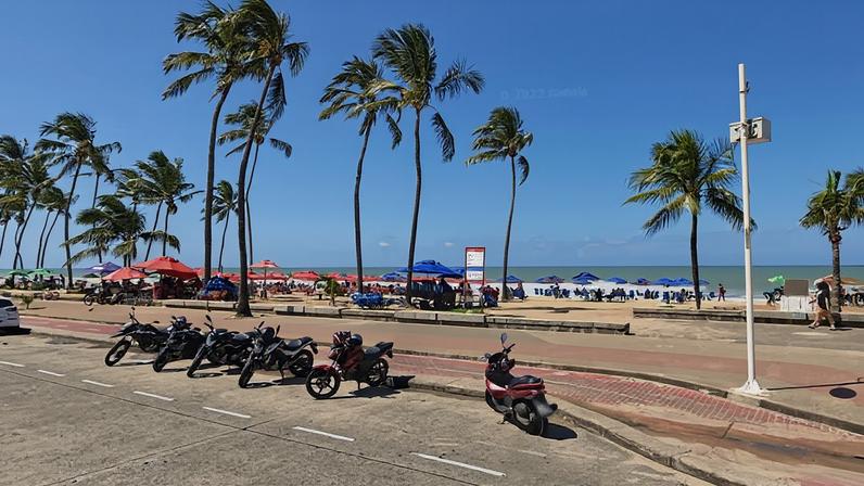 Seis motos e um carro estacionados em via próxima à orla de praia com palmeiras altas. Pessoas sob guarda-sóis coloridos na areia e mar ao fundo sob céu azul com poucas nuvens.
