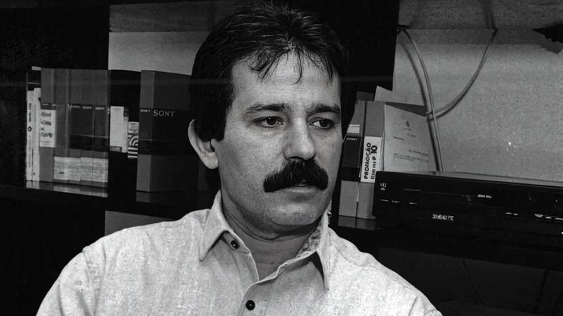 Homem com bigode sentado em cadeira, vestindo camisa clara e calça clara, em sala com dois monitores CRT e equipamentos eletrônicos ao fundo.