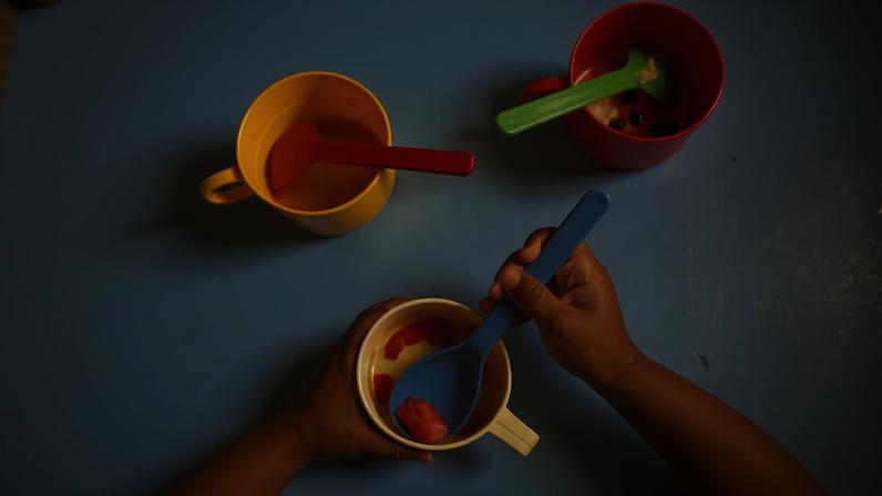 Mãos de criança seguram colher azul e copo branco com pedaços de frutas vermelhas. Sobre mesa azul, há dois copos com colheres: amarelo com colher vermelha e vermelho com colher verde.