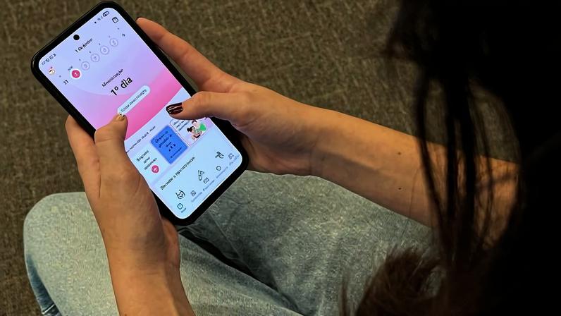 Pessoa sentada com calça jeans clara segura celular com aplicativo aberto que mostra contagem regressiva de 11 dias. Fundo neutro e foco nas mãos e tela do aparelho.