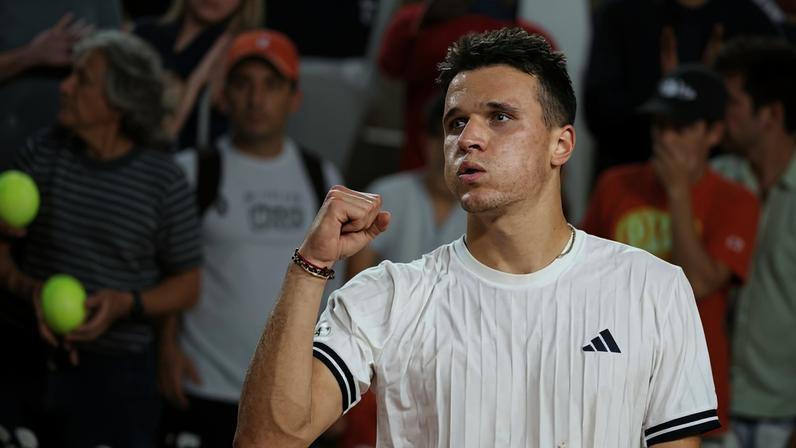 Jogador de tênis veste camiseta branca com logo da Adidas e faz gesto de comemoração com punho cerrado. Público desfocado ao fundo observa a partida.