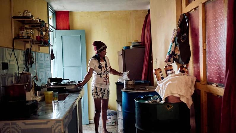 Mulher de camiseta branca e shorts está em cozinha modesta com paredes amarelas. Ela segura um saco plástico perto de um fogão e barris azuis. Porta azul ao fundo e utensílios pendurados na parede.