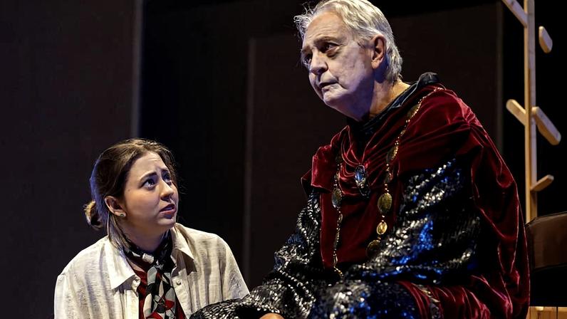 Homem idoso com coroa dourada e traje vermelho com mangas de malha metálica está de braços cruzados sob luz de palco, com fundo escuro e fumaça.