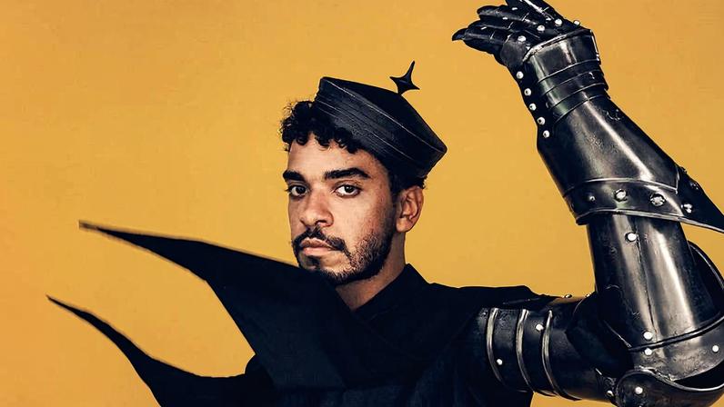 Homem de pele morena veste roupa preta e armadura metálica nos braços, com uma das mãos levantada. Usa chapéu preto com formato pontiagudo contra fundo amarelo.