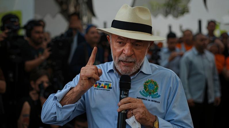 Lula usa chapéu branco e camisa azul clara, segura microfone e gesticula com a mão direita durante discurso. Ao fundo, várias pessoas observam e algumas filmam o evento.