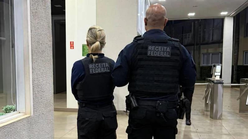 Dois agentes da Receita Federal, um homem e uma mulher, estão de costas em um corredor interno iluminado. Ambos vestem coletes pretos com a inscrição 'Receita Federal' nas costas. O ambiente tem piso claro e paredes brancas, com outras pessoas ao fundo.