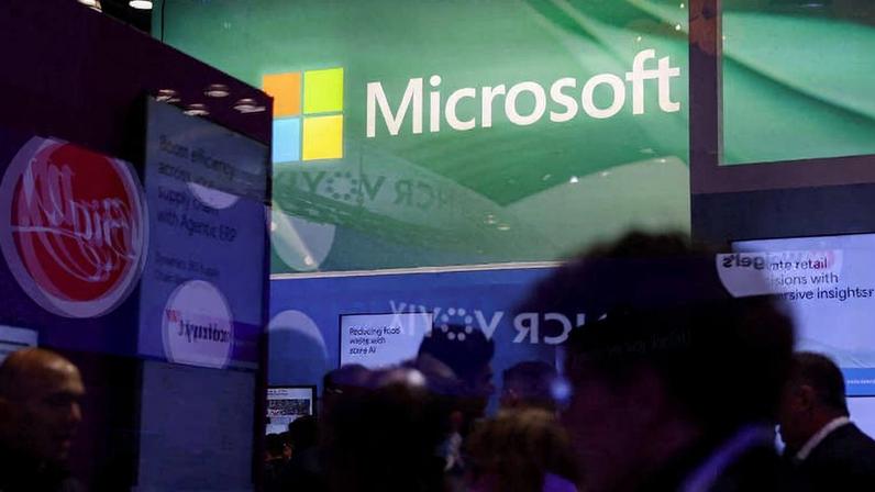 Pessoas em frente a um estande iluminado com o logo da Microsoft em destaque no fundo. Telas e painéis exibem conteúdos digitais, com ambiente de feira ou conferência tecnológica.