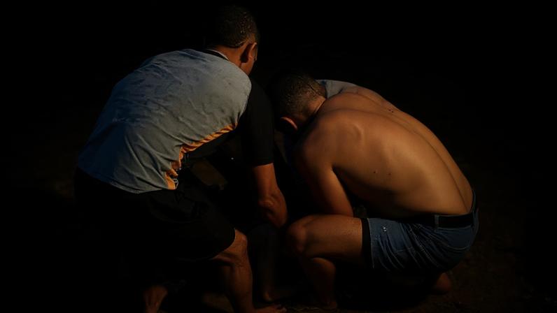 Duas pessoas agachadas ajudam uma mulher deitada na margem de um rio. A mulher está deitada de barriga para baixo na areia, enquanto os dois homens seguram seus braços e ombros.
