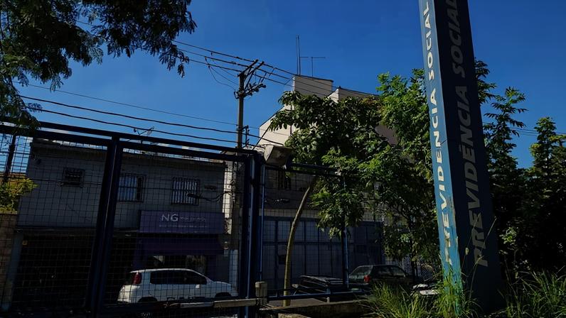Fotografia tirada durante o dia mostra a entrada de uma agência da Previdência Social. Em primeiro plano, há um portão de grade metálica azul e, à direita, um totem vertical azul-escuro com a inscrição “PREVIDÊNCIA SOCIAL” em letras brancas. Atrás da grade, vê-se um pequeno estacionamento com carros parados e um prédio de dois andares. Do lado esquerdo, uma fachada comercial com toldo roxo aparece parcialmente ao fundo. Árvores com folhagem verde cercam o local e projetam sombras sobre a cena. O céu está limpo e azul, com poucas nuvens brancas. A imagem transmite a aparência de um órgão público em funcionamento em um dia ensolarado.