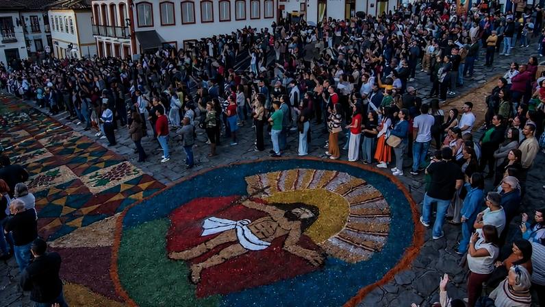 Multidão reunida em rua de paralelepípedos com casas coloniais ao redor, observando tapete colorido no chão que representa figura religiosa com auréola. Céu azul com poucas nuvens ao fundo.
