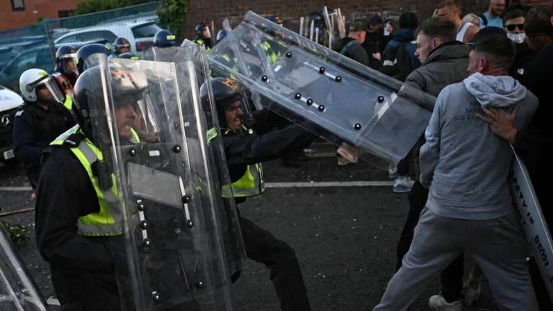 Policiais com escudos transparentes e capacetes enfrentam manifestantes em rua. Manifestantes empurram escudos enquanto outros observam ao fundo.