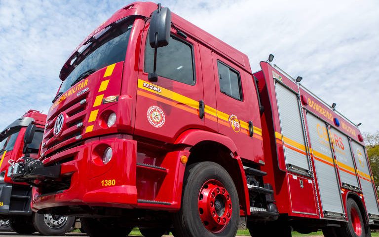 Vários caminhões de bombeiros vermelhos alinhados em fila sobre pavimento, com céu parcialmente nublado ao fundo. Cada veículo exibe faixas amarelas e vermelhas, além de símbolos oficiais dos bombeiros.
