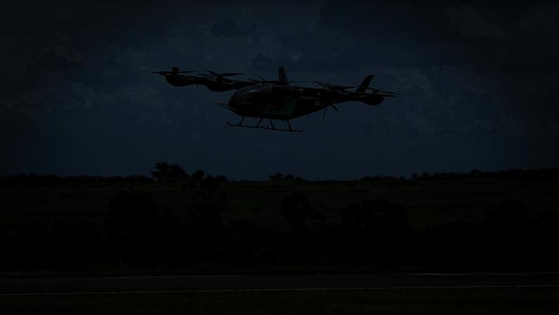 Drone de transporte preto com hélices visíveis voa baixo sobre pista asfaltada, com vegetação verde e céu nublado ao fundo.