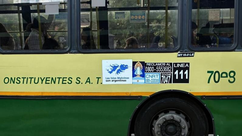 Ônibus verde e amarelo com pneu dianteiro esquerdo furado parado em via asfaltada. Passageiros são visíveis pelas janelas superiores.