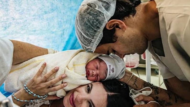 Bebê recém-nascido enrolado em manta branca com touca, deitado sobre o peito da mãe na cama do hospital. Pai inclina a cabeça tocando a do bebê, ambos próximos e em ambiente hospitalar.