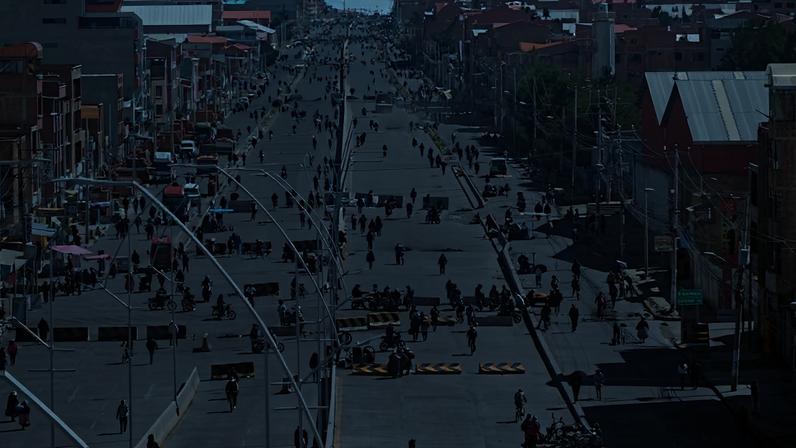 Rua larga e longa em área urbana com muitos pedestres e ciclistas circulando. Edifícios residenciais e comerciais alinham ambos os lados da via sob céu parcialmente nublado.