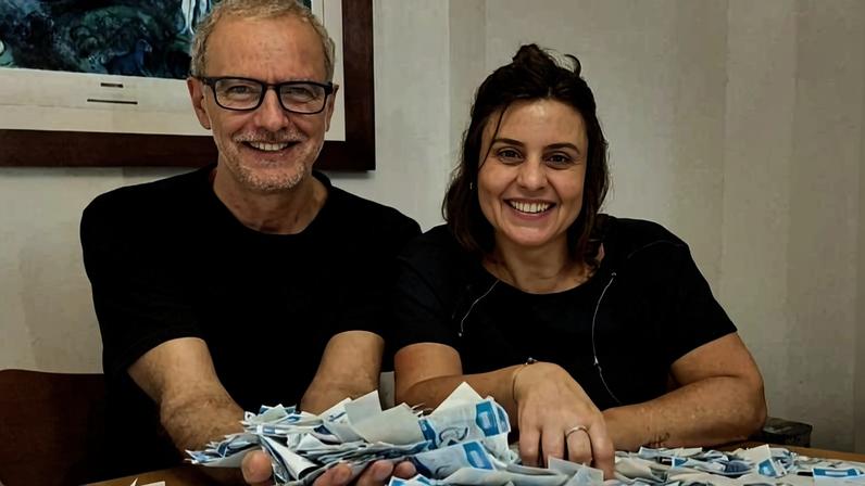Homem e mulher sentados lado a lado em mesa coberta por muitos papéis dobrados. Ambos sorriem para a câmera em ambiente interno com quadro na parede ao fundo.