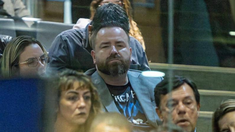 Homem de terno cinza e camiseta preta sentado entre outras pessoas em ambiente fechado, com expressão neutra. Pessoas ao redor estão focadas no evento, algumas com óculos, em fileiras de assentos.