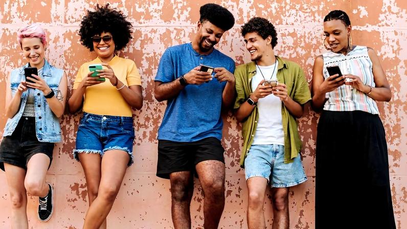 Cinco jovens encostados em parede texturizada cor de ferrugem, todos olhando para seus celulares. Eles vestem roupas casuais de verão, com shorts, camiseta, saia longa e colete jeans.