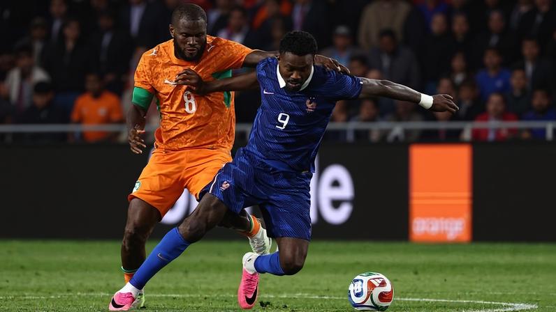 Jogador da França com camisa azul número 9 tenta controlar a bola enquanto é marcado por jogador da Costa do Marfim com camisa laranja número 8 em campo de futebol durante partida noturna.
