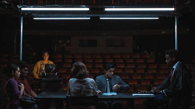 Cinco pessoas sentadas ao redor de uma mesa retangular em sala escura, iluminadas por luzes fluorescentes acima. Quatro estão sentadas, uma em pé ao fundo com um objeto na mão. Ambiente sugere interrogatório ou reunião formal, com cadeiras vermelhas ao fundo.