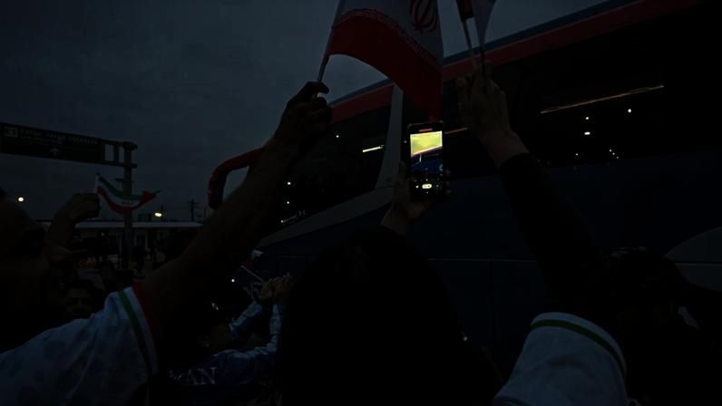 Grupo de pessoas acena bandeiras do Irã e grava com celular próximo a ônibus azul e branco em área externa sob céu nublado.