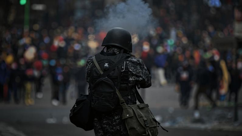 Militar com uniforme camuflado e capacete dispara arma de fogo em direção a multidão desfocada em ambiente urbano.