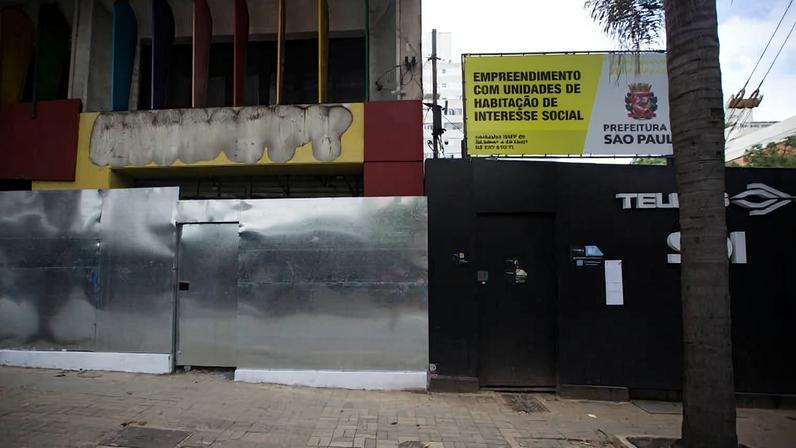 Fachada de prédio com tapumes metálicos prateados e porta preta. Acima, placa amarela da Prefeitura de São Paulo anuncia empreendimento com unidades de habitação de interesse social. Calçada com árvore e poste visíveis em frente.
