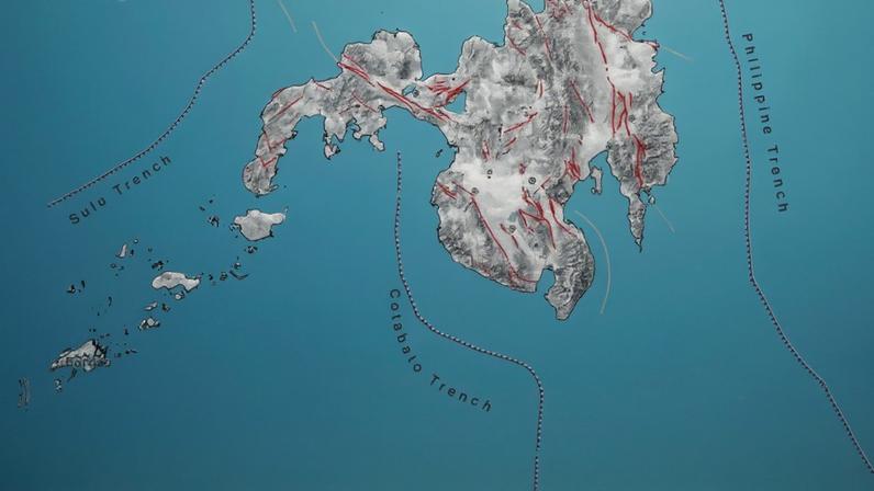 Mapa mostra a ilha de Mindanao nas Filipinas com cidades marcadas, incluindo Davao, Cagayan de Oro e Zamboanga. Linhas pontilhadas indicam as trincheiras de Sulu e Cotabato no mar ao redor. Um ponto vermelho destaca uma localização ao sul da ilha, próxima à Trincheira de Cotabato.