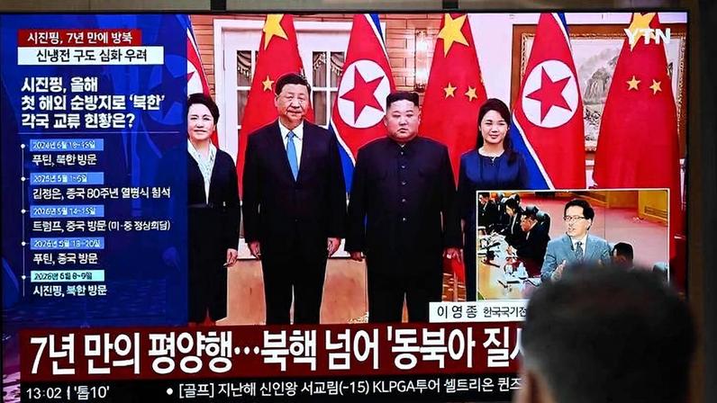 Líderes da Coreia do Norte e China posam juntos em sala oficial com bandeiras dos dois países ao fundo. Tela de TV exibe notícias em coreano sobre visita e relações bilaterais.