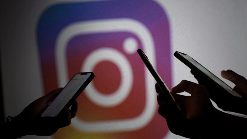 Duas mãos seguram celulares com telas iluminadas em primeiro plano, com o logo colorido do Instagram desfocado ao fundo.