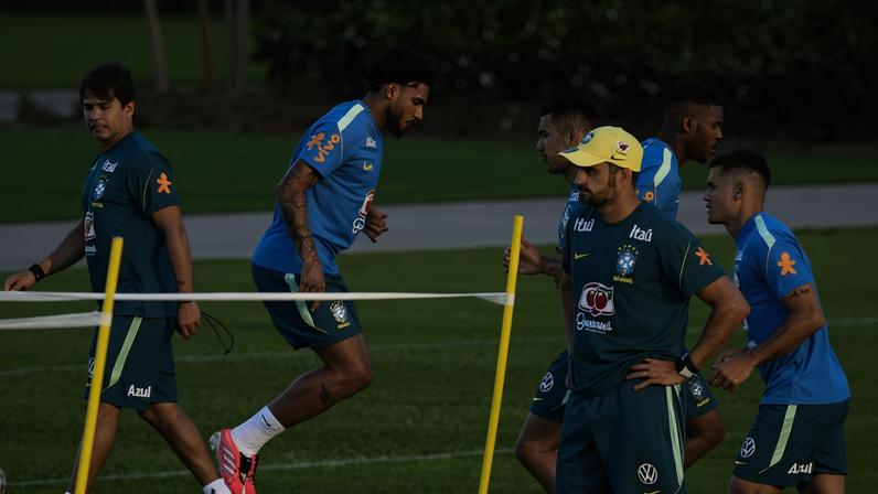 Seis jogadores da seleção brasileira de futebol participam de treino em campo gramado. Um deles está no centro saltando sobre uma corda baixa, enquanto os demais observam e se preparam para o exercício. Todos vestem uniformes oficiais de treino com as cores azul e verde, e o ambiente é ao ar livre com vegetação ao fundo.