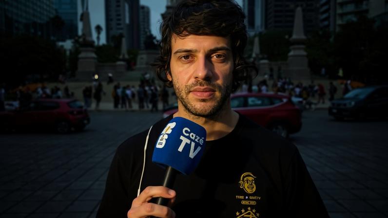 Homem jovem segura microfone azul da Cazé TV em praça movimentada com monumento e prédios ao fundo. Pessoas e carros circulam na área aberta.