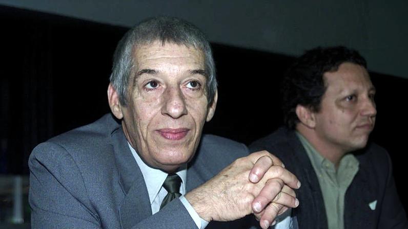Homem de cabelos grisalhos curtos veste terno cinza, camisa clara e gravata escura, com as mãos entrelaçadas à frente. Ao fundo, homem de cabelo escuro e camisa verde aparece de perfil.