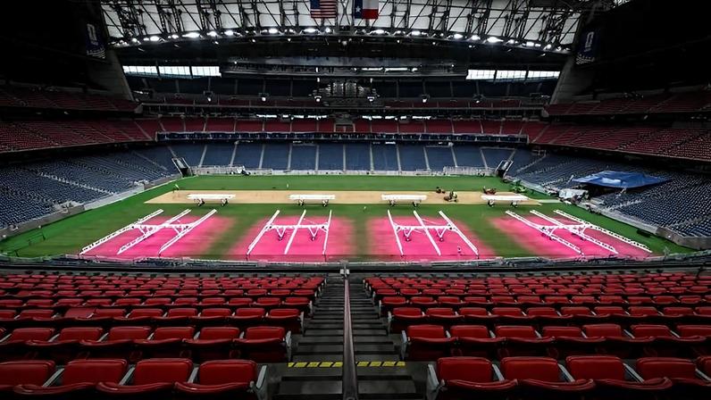 Estádio vazio com arquibancadas vermelhas e azuis. No campo, áreas iluminadas em rosa com marcações para salto em altura. Estrutura metálica e bandeiras dos EUA e Texas no teto.