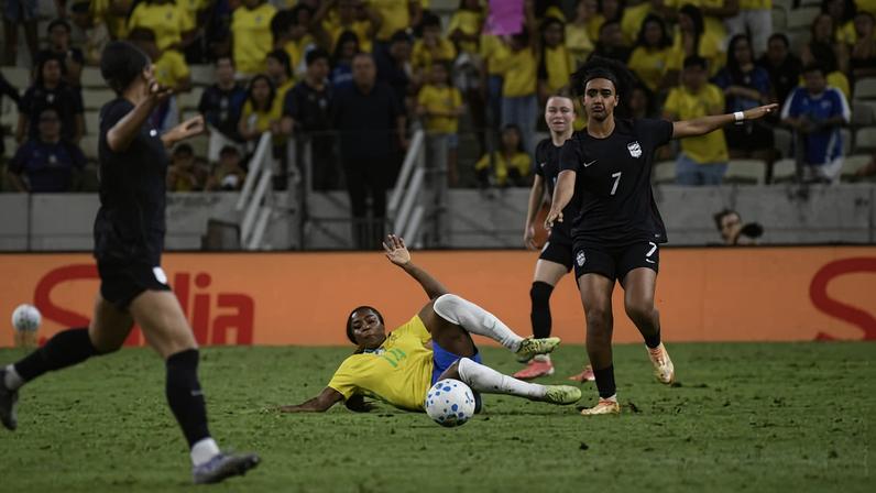 Jogadora da seleção brasileira de futebol feminino executa carrinho para disputar a bola com adversária vestindo uniforme preto durante jogo em estádio com público ao fundo.