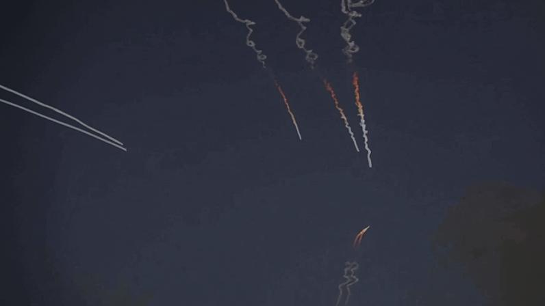 Três foguetes deixam rastros de fumaça e fogo ascendendo em direção ao céu azul escuro. À esquerda, um avião deixa um rastro branco horizontal.