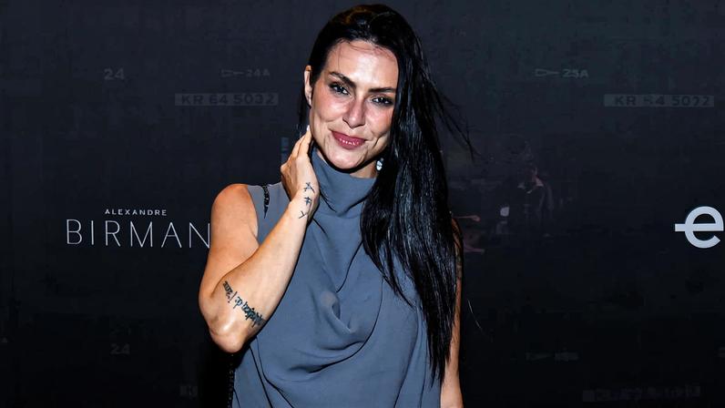 Mulher de cabelos longos e pretos usa vestido cinza sem mangas e está em pé diante de fundo preto com logos e textos brancos, incluindo '35 anos', 'elo' e 'Conspiração'.