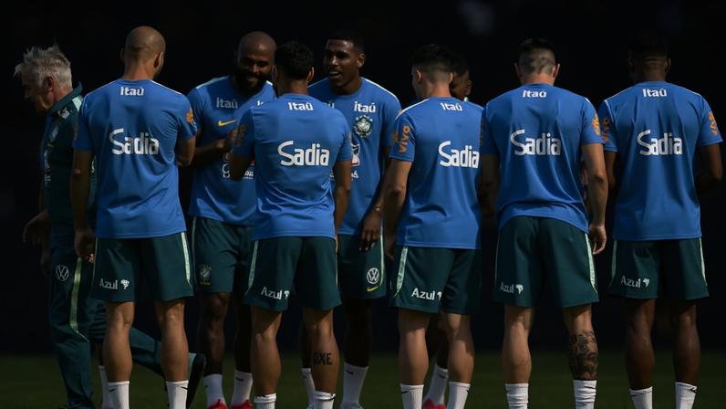 Dez jogadores da seleção brasileira de futebol vestindo uniformes azuis e verdes estão reunidos em círculo no campo de treino, conversando e interagindo entre si. Um técnico de cabelos grisalhos está à esquerda, observando o grupo. O gramado verde e equipamentos esportivos aparecem ao fundo.