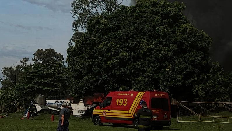 Casa residencial em chamas com fumaça preta saindo do telhado. Bombeiros com uniformes vermelhos e amarelos utilizam mangueiras para apagar o fogo. Árvores e outras casas aparecem ao redor, céu parcialmente nublado.