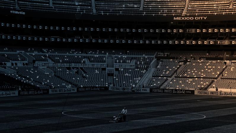 Funcionário empurra cortador de grama no centro do campo de futebol vazio no estádio Azteca, com arquibancadas vermelhas e brancas ao fundo. Faixas com a frase 'Football Unites the World' e bandeiras decoram o local.